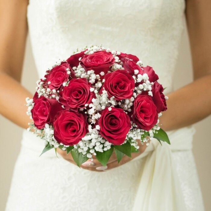 Set against the serene backdrops familiar to Harlington, this intimate, chest-level portrait reveals the artistry behind a traditional wedding bouquet. Immaculately arranged, the bouquet features robust, jewel-toned red roses, each bloom nested side by side to create a lush, domed centrepiece. The roses are fresh and alive, ranging from just-budding to fully open, their tightly furled petals glowing under soft, natural light similar to that in a quiet Harlington morning. White gypsophila, or baby's breath, sparkles among the roses with a cloud-like softness, a classic accent that introduces lightness and textural contrast. Sprigs of glossy green leaves gently support the bouquet at its base, reminiscent of the lush hedgerows along Sipson Road. The graceful, manicured hands of the bride enfold the handle, while her ivory wedding dress, detailed in floral lace and delicate embroidery, peeks above the blooms. A flowing, gentle sash completes the look, trailing like silk in the breeze. The neutral, blurred background keeps focus locked onto the romantic composition. Ideal for exploring the character of Harlington-inspired weddings or bridal fashion, this bouquet invites thoughts of ceremony, hope, and heartfelt celebration.
